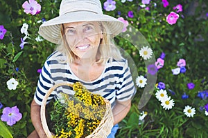 Happy smiling elderly senior woman having fun posing in summer garden with flowers in straw hat. Farming, gardening, agriculture,