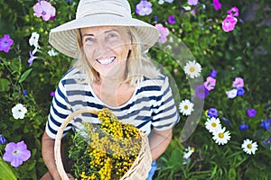 Happy smiling elderly senior woman having fun posing in summer garden with flowers in straw hat. Farming, gardening, agriculture,