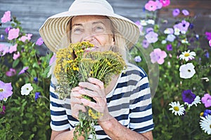 Happy smiling elderly senior woman having fun posing in summer garden with flowers in straw hat. Farming, gardening, agriculture,