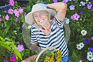 Happy smiling elderly senior woman having fun posing in summer garden with flowers in straw hat. Farming, gardening, agriculture,