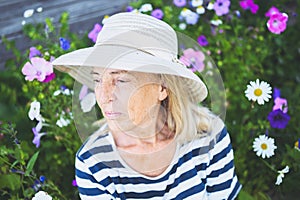 Happy smiling elderly senior woman having fun posing in summer garden with flowers in straw hat. Farming, gardening, agriculture,