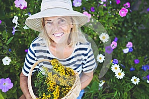 Happy smiling elderly senior woman having fun posing in summer garden with flowers in straw hat. Farming, gardening, agriculture,