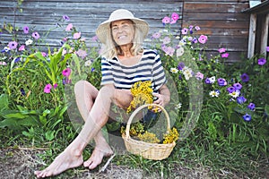 Happy smiling elderly senior woman having fun posing in summer garden with flowers in straw hat. Farming, gardening, agriculture,