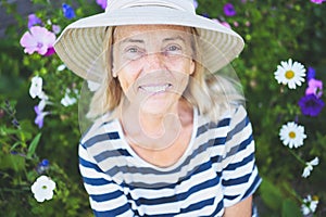 Happy smiling elderly senior woman having fun posing in summer garden with flowers in straw hat. Farming, gardening, agriculture,