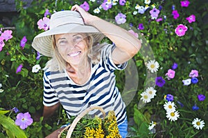 Happy smiling elderly senior woman having fun posing in summer garden with flowers in straw hat. Farming, gardening, agriculture,