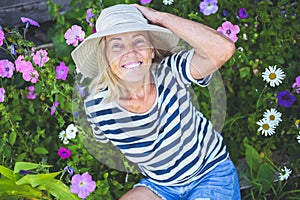 Happy smiling elderly senior woman having fun posing in summer garden with flowers in straw hat. Farming, gardening, agriculture,