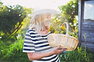 Happy smiling elderly senior woman in straw hat having fun posing in summer garden with flowers in basket. Farming, gardening,