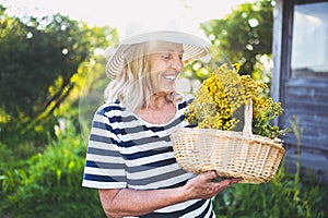 Happy smiling elderly senior woman in straw hat having fun posing in summer garden with flowers in basket. Farming, gardening,