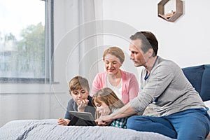 Happy Smiling Family Sitting On Bed Use Tablet Computer, Parents Spending Time With Son And Daughter