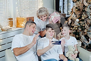 Happy smiling family sitting next to a beautifully decorated Christmas tree, greeting and showing gifts over the phone