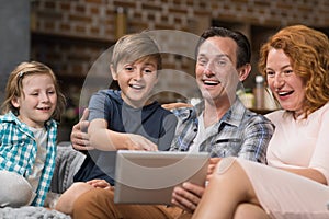 Happy Smiling Family Use Tablet Computer Sitting On Couch In Living Room, Parents Spending Time With Son And Daughter