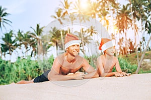 Happy smiling father and son in Santa hats have sun bath on sand