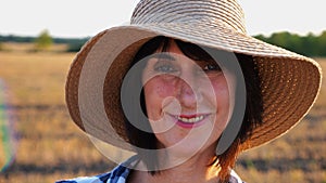 Happy smiling female farmer looks into camera standing at wheat field. Portrait of adult beautiful agronomist with straw