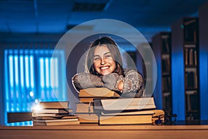 Happy smiling female student has completed the exam preparation. The girl leaned on a stack of books sitting in the library at