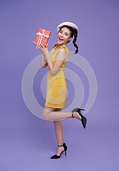 happy smiling fun asian girl in yellow dress holding present box raise up leg isolated over purple background