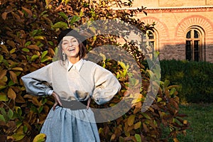 Happy smiling girl in black beret and grey dress stands near the autumn tree, old brick building on the background