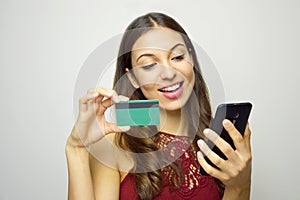 Happy smiling girl holding and looking to her smart phone with credit card in other hand on white background. E-commerce woman.