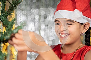 Happy smiling girl kid in santa hat decorating Christmas tree for festive holyday celebration at home. photo
