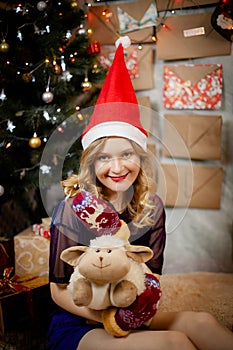 Happy smiling girl in red christmas hat and mittens with gifts and toy near decorated christmas tree