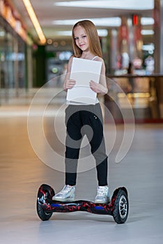 Happy and smiling girl rides on mini segway at trading mall. Teenager riding on hover board or gyroscooter and holding blank sheet