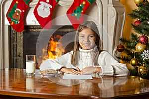 Happy smiling girl sitting by fireplace and writing letter to Sa