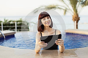 Happy smiling girl using digital tablet computer while bathing in a swimming pool