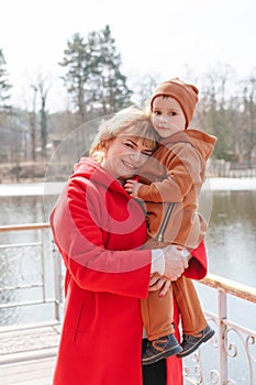 Happy smiling grandmother holds her beloved grandson in her arms. Family portrait on a walk in the forest near the lake.