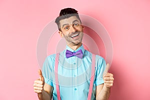 Happy smiling guy in bow-tie and suspenders showing thumbs up, praising good job, approve nice work, standing over pink