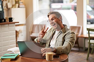 Happy smiling guy in glasses, sits in cafe, shows peace sign at laptop camera, video chats, connects to online meeting