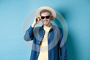 Happy smiling guy going on summer vacation, wearing straw hat and black sunglasses, standing on blue background