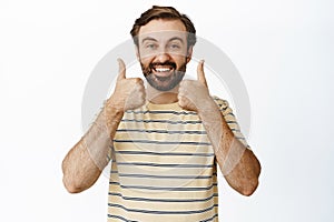 Happy smiling guy shows his support with thumbs up near face, likes and approves smth, stands against white background