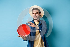 Happy smiling guy throwing frisbee, looking aside at friend, standing on blue background in straw hat