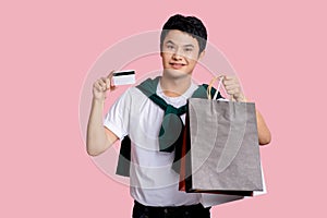 Happy smiling handsome Asian man carrying shopping bags and showing credit card in light pink isolated studio background