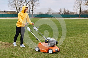 Happy smiling handsome man wearing yellow hoodie and black pants, trimming grass on field with grass-cutter, worker mow lawn with