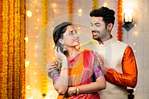 Happy smiling indian couple with traditional ethnic wear looking each other during diwali festival at home - concept of