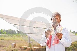 happy smiling indian farmer couting money while standing in front of solar panel at farmland - concept of electric power