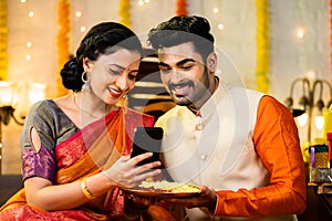 Happy smiling indian wife showing mobile phone to husband by holding flower plate during diwali festival at home -