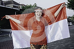 Happy smiling Indonesian woman wearing red kebaya holding Indonesia's flag to celebrate Indonesia Independence Day. Outdoor