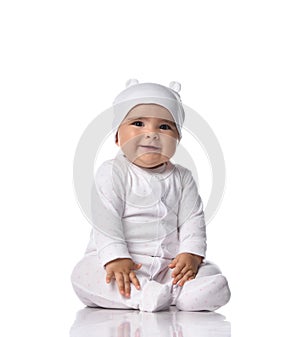 Happy smiling infant baby toddler sits on the floor in white onepiece jumpsuit overall and funny hat with ears