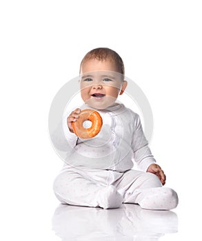Happy smiling infant baby toddler in white onepiece jumpsuit overall sits on the floor with donut in hands gives it us