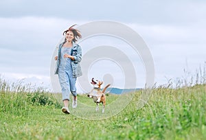 Happy smiling jogging female with fluttering hairs and her beagle dog running and looking at eyes. Walking by meadow grass path in