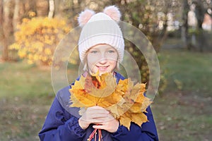 Happy smiling kid girl in a warm knitted hat collects bouquet of yellow leaves. Autumn in the park.