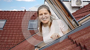 Happy smiling and laughing woman looking out of the open attic window in her apartment under the red tiled roof