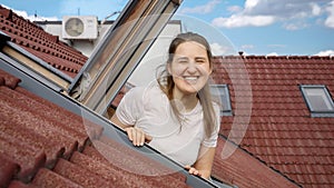 Happy smiling and laughing woman looking out of the open attic window and waves hand in camera