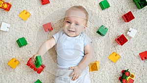 Happy smiling little baby boy lying on carpet covered with colorful toys, blocks and bricks. Concept of children