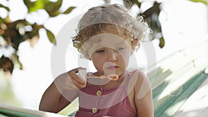 Happy smiling little baby girl has fun blowing soap bubbles sitting on the hammock in the sunny day in green home garden, blue-