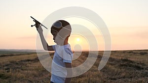 Happy smiling little boy child in helmet aviator pilot of airplane playing with toy wooden plane in field