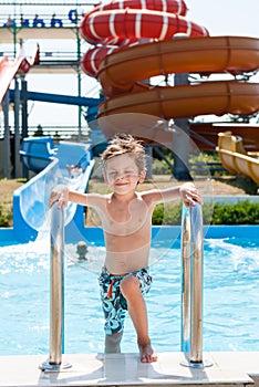 Happy smiling little boy in shorts standing near swimming pool in water park with water tube slides