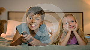 Happy smiling little caucasian children boy girl brother sister watching TV together lying on bed in bedroom kids
