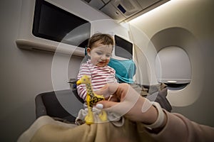 Happy smiling little cute toddler sitting in the baby bassinet of an airplane. laughs and looks at her mother`s hand